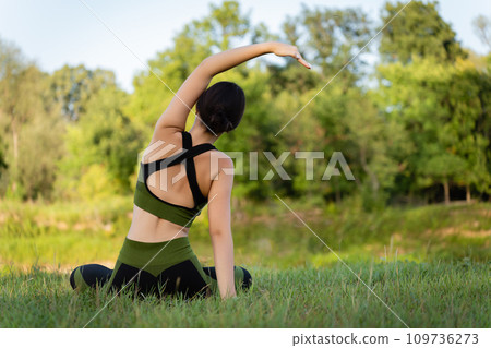 View from the back. Sitting on the grass, a teenager girl performs tilts to the sides with a beautiful view of the summer nature. Physical form, harmony View from the back. Sitting on the grass, a teenager girl performs tilts to the sides with a beautiful view of the summer nature. Physical form, harmony 109736273