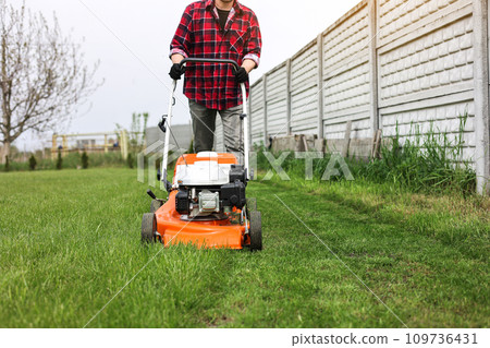 view of man in casual clothes mows lawn with lawn mower at backyard of his house. Husband takes care of garden on spring cloudy day. Modern gasoline garden equipment. Landscaping work 109736431