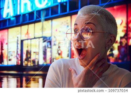 Shocked and excited young woman standing on evening street, looking on shopping mall windows showing big sales. Eyeglass reflection Shocked and excited young woman standing on evening street, looking on shopping mall windows showing big sales. Eyeglass reflection 109737034