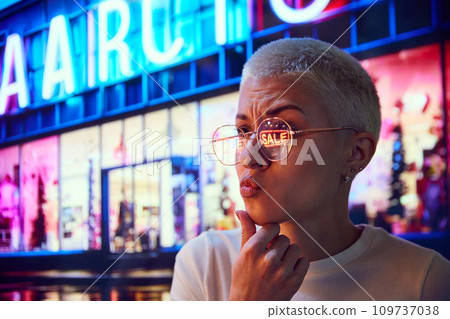 Young woman standing on evening street, looking on shopping mall windows with interest. Big sales. Eyeglass reflection Young woman standing on evening street, looking on shopping mall windows with interest. Big sales. Eyeglass reflection 109737038