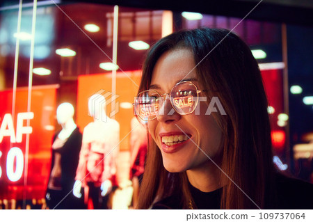Close-up portrait of smiling young woman with reflection on glasses looking on shop windows. Big sales in shopping mall. Close-up portrait of smiling young woman with reflection on glasses looking on shop windows. Big sales in shopping mall. 109737064