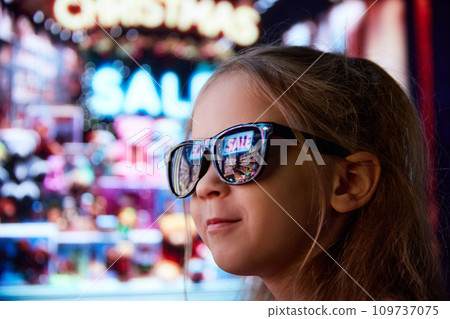 Close-up image of little girl in sunglasses standing on street and looking at shop window. Lights reflection on glasses. Big sales Close-up image of little girl in sunglasses standing on street and looking at shop window. Lights reflection on glasses. Big sales 109737075