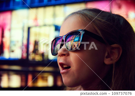 Close-up image of little girl in sunglasses standing on evening street and looking at shopping mall. Lights reflection on glasses Close-up image of little girl in sunglasses standing on evening street and looking at shopping mall. Lights reflection on glasses 109737076