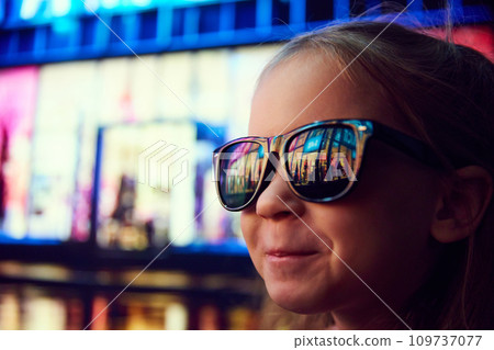Close-up image of little girl in sunglasses standing on evening street and looking at shopping mall. Lights reflection on glasses Close-up image of little girl in sunglasses standing on evening street and looking at shopping mall. Lights reflection on glasses 109737077