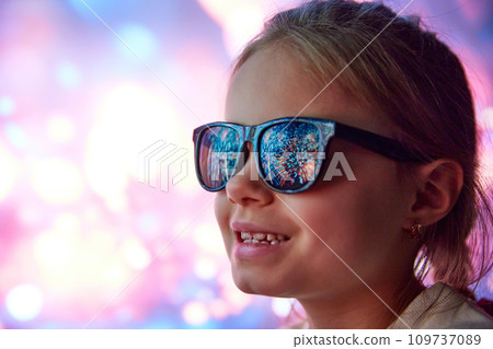Happy smiling little girl, cheerful child in sunglasses looking on fireworks. Reflection on glasses. Outdoor holidays celebration 109737089