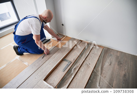 Man in work overalls using tape measure while installing laminate flooring in apartment under renovation. Male builder checking distance from wall to laminate board. Hardwood floor renovation concept. 109738308