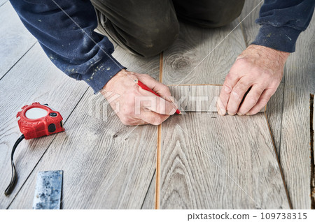 Close up of man construction worker with pencil in hand drawing line on laminate timber board. Male worker preparing laminate material for floor installation at home during renovation. Close up of man construction worker with pencil in hand drawing line on laminate timber board. Male worker preparing laminate material for floor installation at home during renovation. 109738315