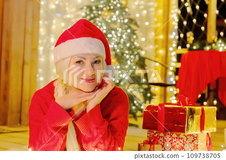 Woman in Santa hat by Christmas tree. Festive setting. Illustrating holiday cheer and celebration. She is posing for a picture, and there are presents nearby, adding to the festive atmosphere. Woman in Santa hat by Christmas tree. Festive setting. Illustrating holiday cheer and celebration. She is posing for a picture, and there are presents nearby, adding to the festive atmosphere. 109738705