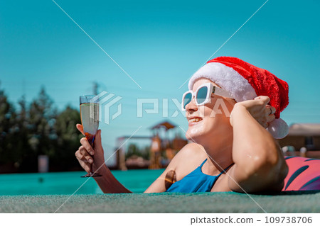 Woman pool Santa hat. A happy woman in a blue bikini, a red and white Santa hat and sunglasses poses near the pool with a glass of champagne standing nearby. Christmas holidays concept. 109738706