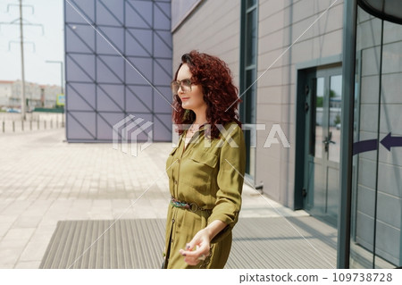 Portrait of a woman standing near a supermarket building. Caucasian model with long brunette hair, wears sunglasses and a khaki dress. 109738728