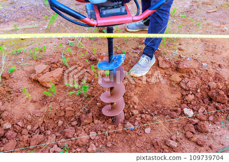 With gasoline drilling machine, worker digs ground in preparation for installation of fence posts 109739704