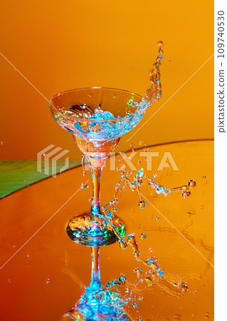 Ice cube falling into glass with martini cocktail against orange background in neon light. Splashes 109740530