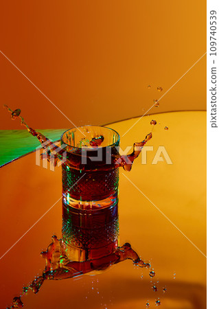 Ice cube falling into glass with strong drink. whiskey and coke against orange background in neon light Ice cube falling into glass with strong drink. whiskey and coke against orange background in neon light 109740539