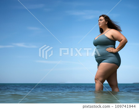 Young chubby woman in gray swimsuit standing in sea water on seashore. Side view. Copy space. 109741944