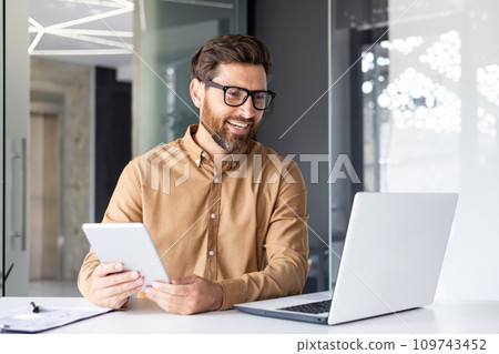 Portrait of a young architect, designer working in the office at a laptop, talking on a video call, holding a tablet in his hands. 109743452