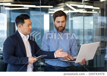 Two young interracial businessmen standing in office, working on laptop, talking and discussing project. 109743479