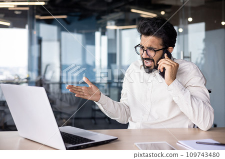 Angry young Indian male businessman sitting in the office at the desk and emotionally talking on the phone. 109743480