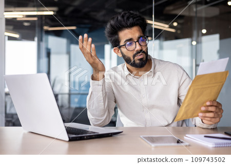 Worried young Indian businessman man sitting in the office at the table and reading the received letter and documents, frustrated and confused throwing up his hands. Worried young Indian businessman man sitting in the office at the table and reading the received letter and documents, frustrated and confused throwing up his hands. 109743502
