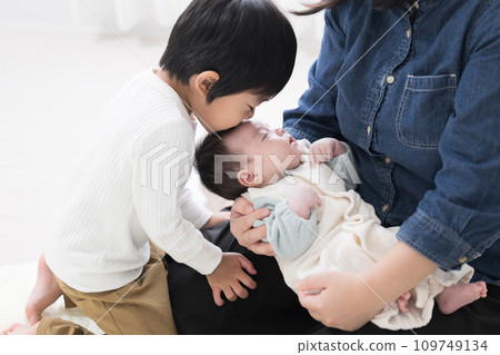 An older brother kisses the forehead of a baby wearing baby clothes sleeping on mom's lap, upper body 109749134