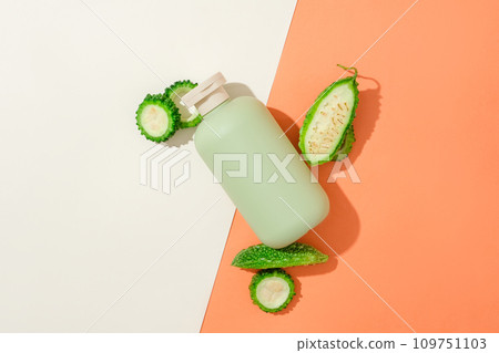 An cosmetics bottle is displayed with bitter melon on an orange-white background. Bitter melon has a lot of water and vitamins, so it has the effect of replenishing moisture and regenerating the skin. 109751103