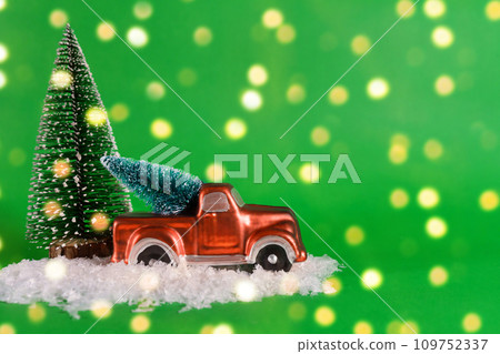 A red pickup truck is carrying a Christmas tree with artificial snow on a green background with bokeh. Banner, New Year card with copy space 109752337