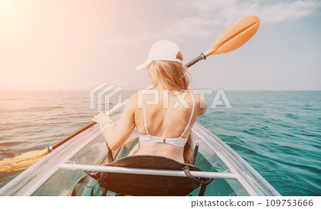 Woman in kayak back view. Happy young woman with long hair floating in transparent kayak on the crystal clear sea. Summer holiday vacation and cheerful female people relaxing having fun on the boat Woman in kayak back view. Happy young woman with long hair floating in transparent kayak on the crystal clear sea. Summer holiday vacation and cheerful female people relaxing having fun on the boat 109753666
