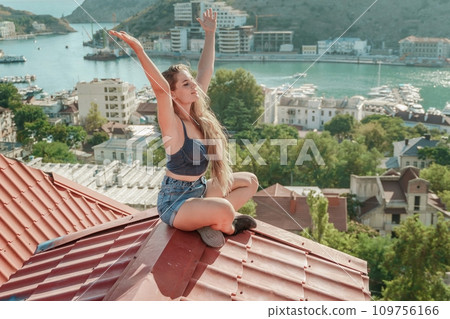 Woman sits on rooftop with outstretched arms, enjoys town view and sea mountains. Peaceful rooftop relaxation. Below her, there is a town with several boats visible in the water Woman sits on rooftop with outstretched arms, enjoys town view and sea mountains. Peaceful rooftop relaxation. Below her, there is a town with several boats visible in the water 109756166