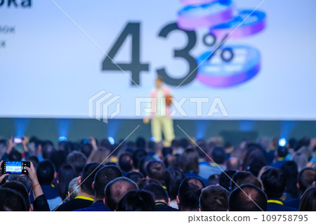 Business crowd listening to male speaker on stage with number and percentage sign being displayed on projection screen at conference hall 109758795