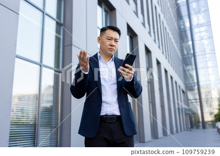 Serious young Asian businessman standing outside office building, holding phone, looking frustrated and worried at screen with arms outstretched. 109759239