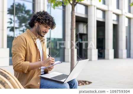 Indian young male student sitting outside on a bench and studying online, watching and listening in headphones to online lectures, writing down in a notebook with concentration. 109759304