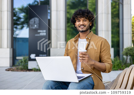 Portrait of a young Indian freelancer man sitting on a bench outside with a laptop, holding a cup of coffee and smiling at the camera. Portrait of a young Indian freelancer man sitting on a bench outside with a laptop, holding a cup of coffee and smiling at the camera. 109759319