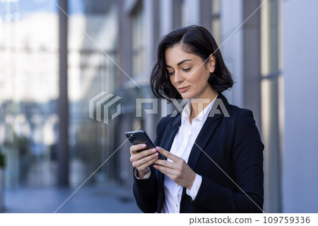 A young business woman is standing near the office center on the street in a business suit and using the phone, typing messages, texting. 109759336