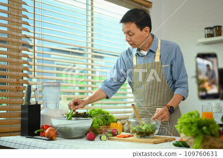 Healthy retired male wearing apron preparing healthy food in kitchen. Healthy Lifestyle concept. Healthy retired male wearing apron preparing healthy food in kitchen. Healthy Lifestyle concept. 109760631