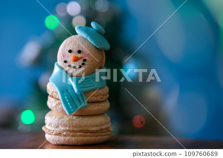 Snowman macaron dessert decorated with hat and scarf against Christmas tree, colorful lights and bokeh background. Merry Christmas and Happy New Year festive concept Snowman macaron dessert decorated with hat and scarf against Christmas tree, colorful lights and bokeh background. Merry Christmas and Happy New Year festive concept 109760689