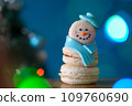 Snowman macaron dessert decorated with hat and scarf against Christmas tree, colorful lights and bokeh background. Merry Christmas and Happy New Year festive concept 109760690