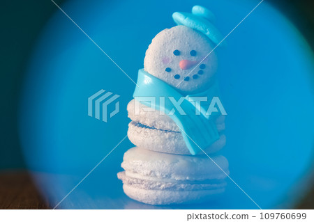 Snowman macaron dessert decorated with hat and scarf inside abstract blue snow globe. Merry Christmas and Happy New Year festive concept Snowman macaron dessert decorated with hat and scarf inside abstract blue snow globe. Merry Christmas and Happy New Year festive concept 109760699