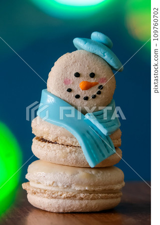 Snowman macaron dessert decorated with hat and scarf against colorful lights and bokeh background. Merry Christmas and Happy New Year festive concept Snowman macaron dessert decorated with hat and scarf against colorful lights and bokeh background. Merry Christmas and Happy New Year festive concept 109760702