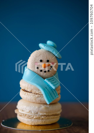 Snowman macaron dessert decorated with hat and scarf against blue background. Merry Christmas and Happy New Year festive concept Snowman macaron dessert decorated with hat and scarf against blue background. Merry Christmas and Happy New Year festive concept 109760704