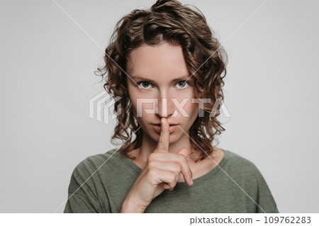 Curly caucasian young woman looks serious, holding finger at her lips asking shut up Curly caucasian young woman looks serious, holding finger at her lips asking shut up 109762283