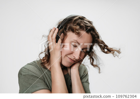 Carefree cheerful young curly woman listen favourite music with hand on her headphones 109762304