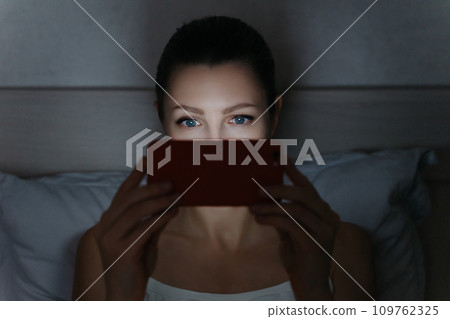Young female using her smartphone late at night in her bed for wathing series instead healthy sleeping. 109762325