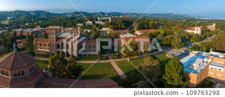 Aerial view of UCLA campus with Gothic tower, red-brick buildings, green lawns, and pathways amidst hilly, tree-covered landscape in soft, golden light. Aerial view of UCLA campus with Gothic tower, red-brick buildings, green lawns, and pathways amidst hilly, tree-covered landscape in soft, golden light. 109763298
