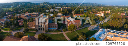 Aerial view of UCLA campus in Westwood, Los Angeles, showcasing Romanesque Revival architecture, modern buildings, green spaces, and a serene city backdrop under a partly cloudy sky. 109763328
