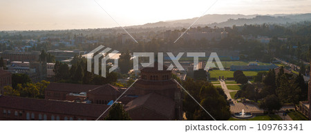 Aerial view of UCLA campus at dawn or dusk with a serene ambiance, featuring a red-brick tower, Neo-Gothic clock tower, sports field, and misty mountains under a vibrant sky. Aerial view of UCLA campus at dawn or dusk with a serene ambiance, featuring a red-brick tower, Neo-Gothic clock tower, sports field, and misty mountains under a vibrant sky. 109763341