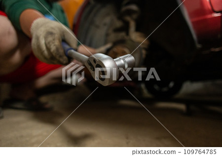 A man repairing a car in a garage holds a ratchet wrench in the cell. A man takes a ratchet wrench to remove rusty bolts from a car wheel hub. Car repair in the garage. 109764785