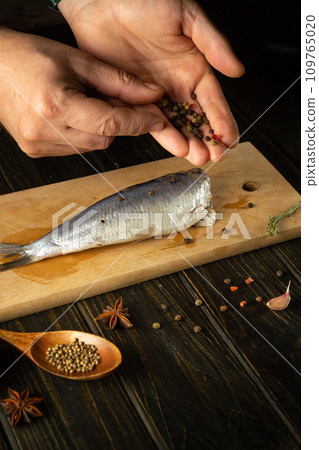 Close-up of a chef's hands adding dry pepper to Clupea fish. Concept of cooking pickled herring with spices on the kitchen table. Close-up of a chef's hands adding dry pepper to Clupea fish. Concept of cooking pickled herring with spices on the kitchen table. 109765020