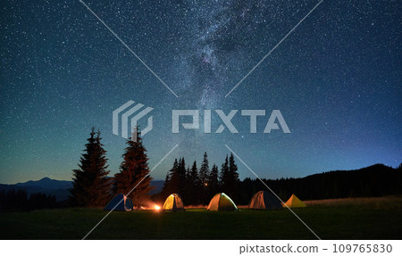 Night camping in mountains under starry sky. Tourist tents in campsite near burning campfire under beautiful sky full of stars with Milky way above forest. Concept of tourism and traveling. 109765830