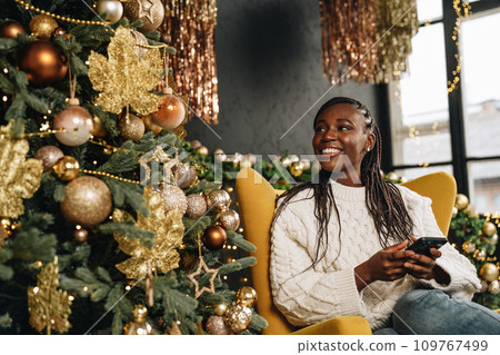 Young beautiful African woman using her phone at home decorated for Christmas season Young beautiful African woman using her phone at home decorated for Christmas season 109767499