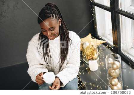 African woman in warm white sweater drinking hot coffee or tea at home 109768053