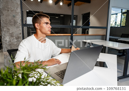 Casual young man in white t-shirt sitting at white desk looking at laptop screen at modern office 109768131
